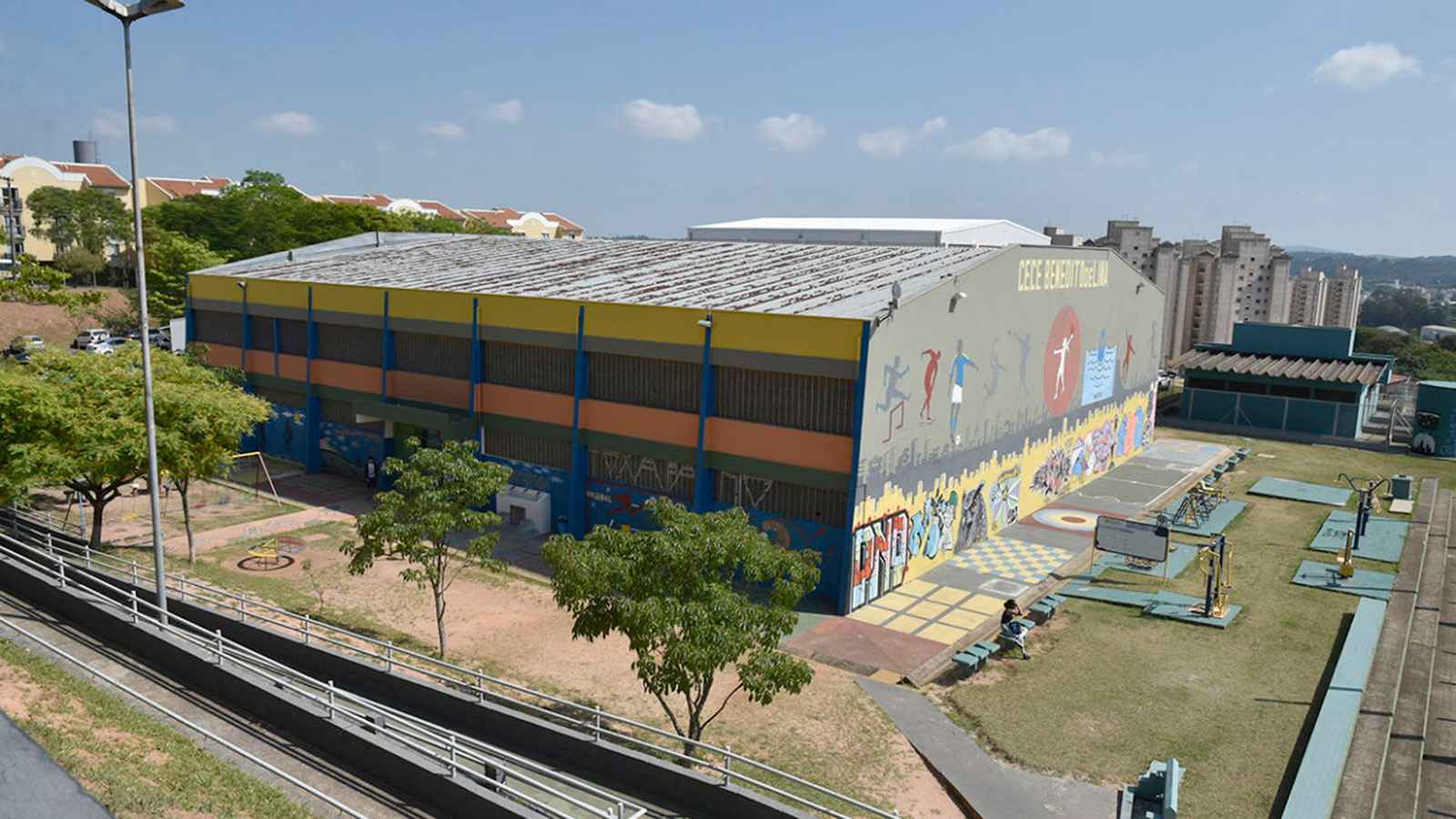 Vista aérea do Centro Esportivo Benedito De Lima em Jundiaí, mostrando o edifício colorido com murais esportivos, áreas de treinamento ao ar livre e a paisagem urbana ao fundo.