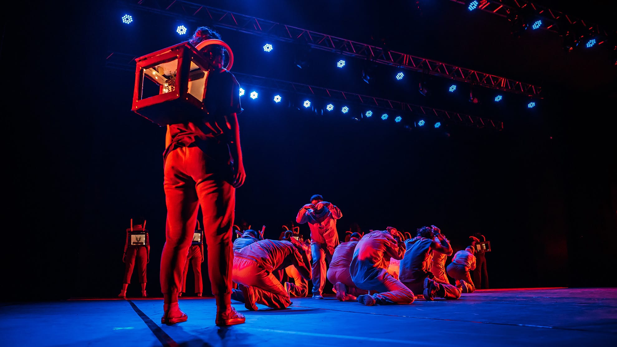 Apresentação no palco durante a mostra competitiva do Hip Hop District de 2022. Uma pessoa em destaque no centro, segurando uma caixa luminosa vermelha com um objeto branco em seu interior. Outros dançarinos em trajes escuros estão posicionados em posturas curvadas e ajoelhadas no chão, evocando uma atmosfera intensa e dramática. O cenário é iluminado por luzes azuis e vermelhas, criando um contraste vibrante entre as silhuetas dos dançarinos e o fundo escuro.