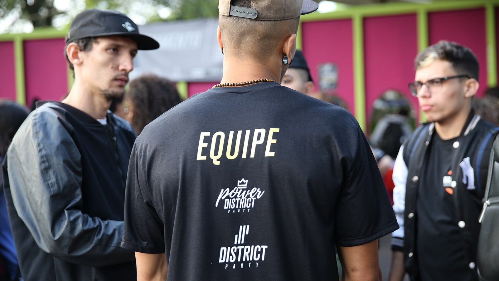 Um membro da equipe do Hip Hop District, vestindo uma camiseta preta com a palavra Equipe, auxiliando dois dançarinos com dúvidas no estande de credenciamento localizado no Parque da Uva.