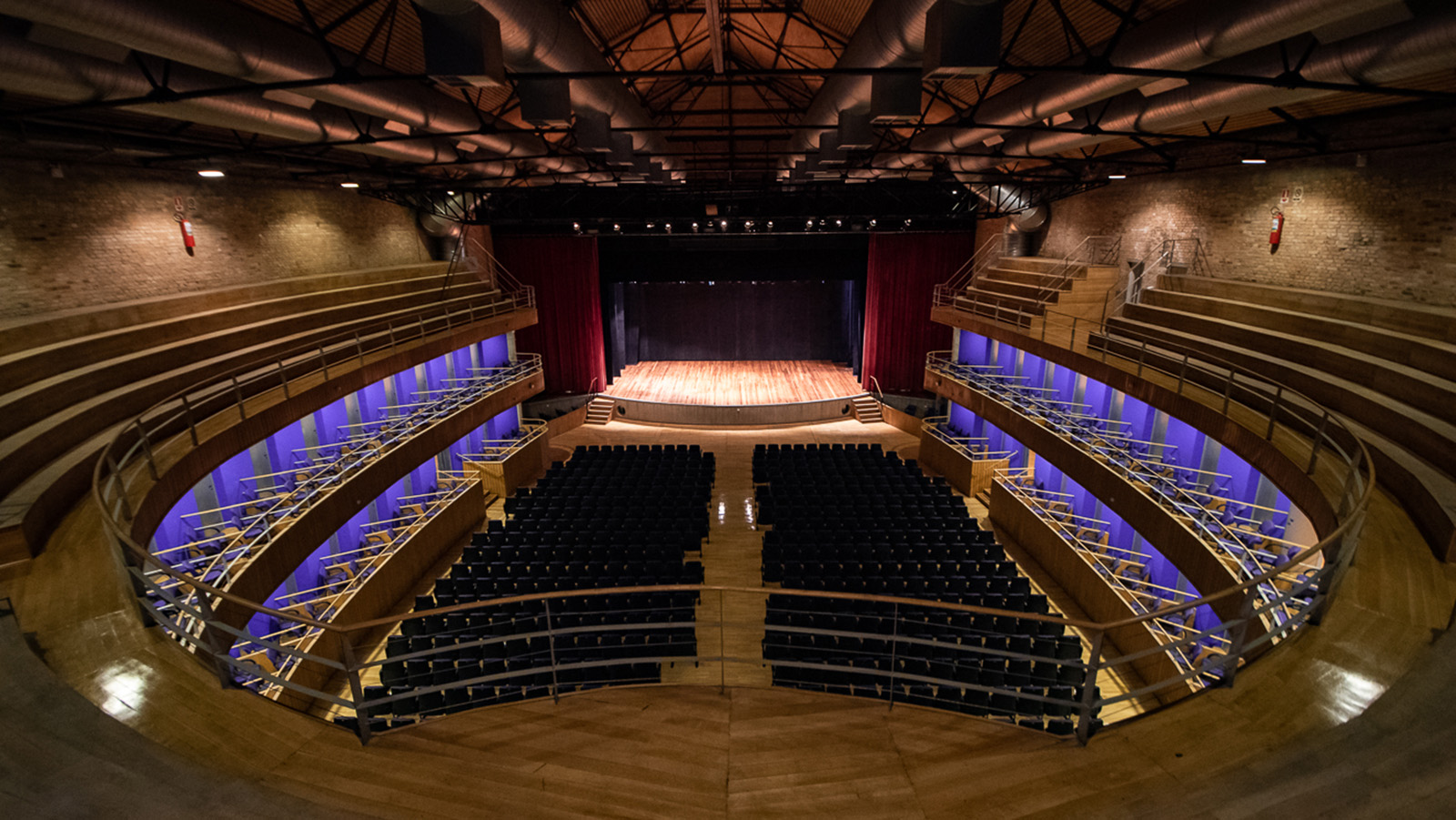 Vista panorâmica do interior do Teatro Polytheama, mostrando os assentos em formato de anfiteatro voltados para um palco de madeira iluminado. As paredes laterais exibem arquitetura moderna com iluminação em tom azul. No alto, a estrutura do teto é composta por vigas metálicas e tubulações. Este é o local onde ocorrerá o evento Hip Hop District.