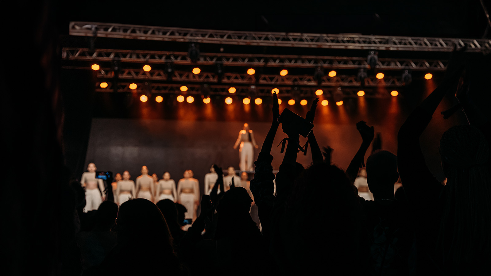 Silhuetas de espectadores em primeiro plano, muitos com os braços erguidos e aplaudindo, e um com uma câmera, celebrando uma apresentação que acaba de ocorrer no evento Hip Hop District. No centro do palco, um(a) artista destaca-se, cercado por um grupo de outros artistas em trajes claros, todos sob uma estrutura de iluminação metálica iluminada por luzes âmbar.